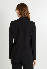 Blazer noir avec une coupe ajustée, présentant un col à revers et des accents de boutons sur les manches. Tissu lisse avec une texture subtile.