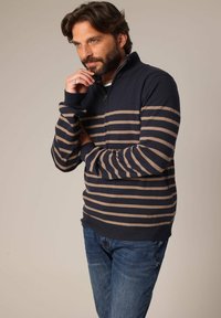 Pull-over rayé bleu marine et beige avec un col zippé mi-hauteur, texture tricotée et poignets côtelés, associé à un jean bleu.