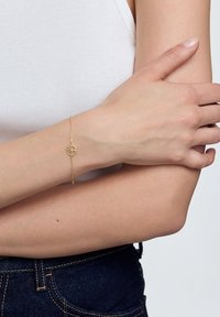 Złota bransoletka z okrągłym zawieszką ozdobioną kwiatowym wzorem, subtelnie połączony łańcuszek, noszona na jasnej skórze, na tle neutralnym.