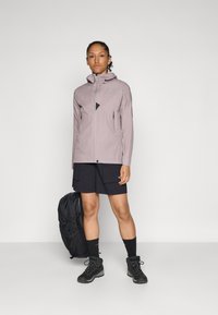 Veste légère et imperméable de couleur rose avec capuche, poches zippées, associée à un short noir, des chaussures de randonnée noires et un sac à dos noir.
