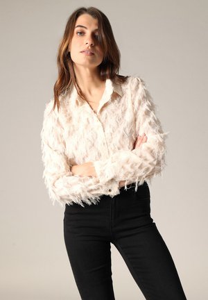 Young woman with brown hair wearing a textured cream blouse and black pants, standing with arms crossed against a plain background.