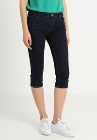 Donkerblauwe denim capri broeken met een aansluitend model, een taille van halverwege de heup en omgeslagen zoom, gecombineerd met witte sneakers en een groen shirt.