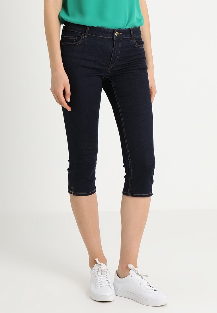 Donkerblauwe denim capri broeken met een aansluitend model, een taille van halverwege de heup en omgeslagen zoom, gecombineerd met witte sneakers en een groen shirt.