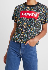 Musta lyhythihainen T-paita, jossa on monivärinen kukkakuvio, ja etupuolella rohkea punainen Levi's-logo. Yhdistetty sinisiin farkkuihin.
