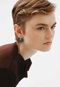 Pendiente circular turquesa con un detalle de aro dorado. El modelo lleva una prenda de color marrón oscuro. El fondo es blanco liso. El diseño limpio enfatiza los colores.