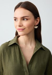 Gouden bloemenoorbellen met witte bloemblaadjes, gedragen met een olijfgroene gekraagde shirt van textiel met een structuur. Eenvoudig ontwerp, minimale accenten.