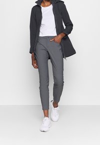 Osoba w ciemnej kurtce, białej koszulce, szarych wąskich spodniach i białych trampkach, idąca z jedną ręką w kieszeni na białym tle.