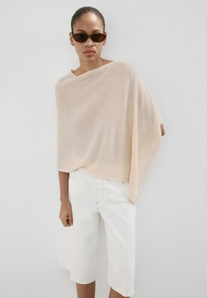 Femme portant un haut ample asymétrique beige clair, un short long blanc et des lunettes de soleil étroites foncées, debout devant un mur clair uni.