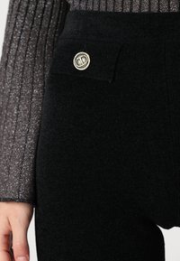 Pantalons en laine noire avec une taille texturée et un détail de bouton métallique sur la poche avant. Fini en tissu doux et lisse.