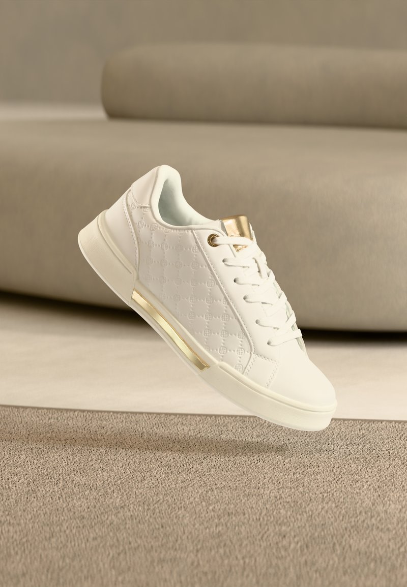 Λευκό αθλητικό παπούτσι με ανάγλυφο σχέδιο, χρυσές λεπτομέρειες στη γλώσσα και τη σόλα, να αιωρείται πάνω από μπεζ χαλί μπροστά από ελαφρύ καναπέ.