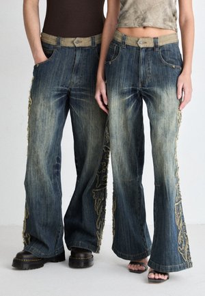 Twee mensen dragen blauw vervaagde wijde jeans met beige geborduurde zijpanelen, gecombineerd met zwarte laarzen en sandalen.