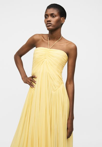 Žena v žlutých bezramínkových plisovaných šatech s halterovým páskem, stojící s jednou rukou na boku před jednoduchým světlým pozadím.