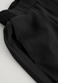 Black fabric pants with a smooth texture, featuring elasticized waistband and loose fit, angled for a close-up view of material and detailing.