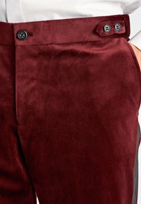 Donkerrode suède broek met zwarte knopen op de tailleband en een zijdelingse verstelriem, gedragen met een witte, ingestopte blouse.