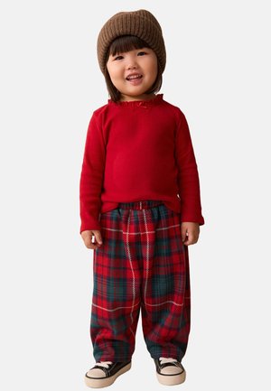 Camicia rossa a maniche lunghe con dettagli a frange abbinata a pantaloni larghi a quadri rossi e verdi. Berretto in maglia marrone e sneakers nere.