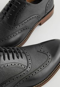 Chaussures habillées en cuir noir avec des accents perforés, surface texturée, design à lacets, et une semelle brune légèrement surélevée.