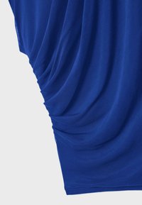 Blauer Stoff mit gerafften, rafﬁnierten Details, die weiche, vertikale Falten gegen einen schlichten hellgrauen Hintergrund bilden.