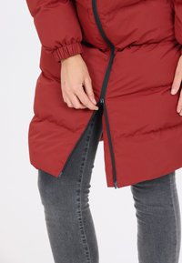 Maroon polstret jakke med elastiske manchetter, en fuldlængde lynlås og en tekstureret overflade. Båret over grå jeans, der viser et nærbillede af lynlåsen.