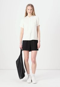 White short-sleeve shirt with a button front, paired with black shorts. Model holds a black tote bag, wearing white sneakers and socks.