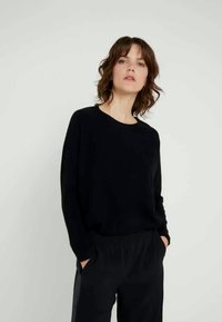 Frau mit lockigem braunem Haar, die ein schwarzes Langarmshirt und schwarze Hosen trägt, steht mit den Händen in den Taschen vor einem einfarbigen hellen Hintergrund.