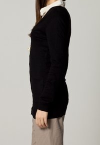 Black knit cardigan with long sleeves, side pockets, and a straight cut, worn over a white collared shirt. Textured fabric appears soft.