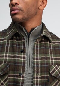 Geruite jas in groen, bruin en crème, met een grijze ritssluiting aan de binnenkant, gestructureerde stof en zwarte knopen aan de voorkant.