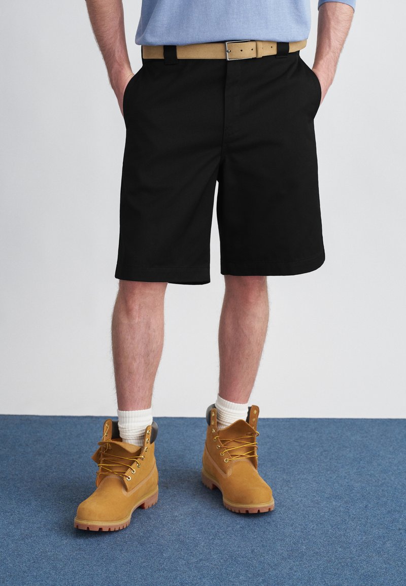 Μαύρο σορτς με πλαϊνές τσέπες, φορεμένο με μπεζ ζώνη. Ταμπά εργασιακά μποτάκια με κίτρινα κορδόνια και υφή στη σόλα, συνδυασμένα με λευκές κάλτσες.