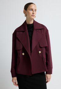 Bordeaux struktur jakke med brede revers, to gyldne løvehovedknapper og forlommer, lagt over en sort bluse.