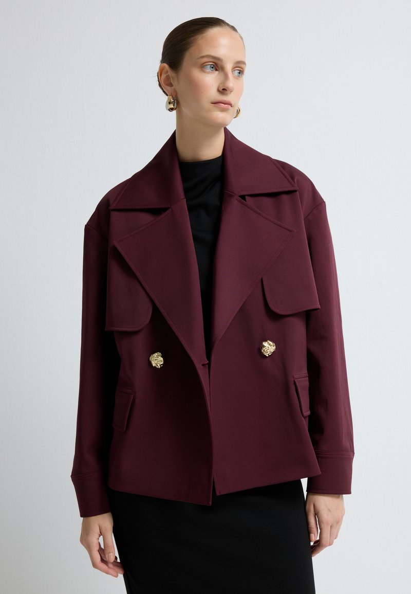 Bordeaux struktur jakke med brede revers, to gyldne løvehovedknapper og forlommer, lagt over en sort bluse.
