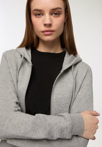 Felpa leggera grigia con zip, dallo stile morbido e dotata di tasche frontali, indossata sopra una maglietta nera. Presenta un cappuccio con cordino e polsini a costine.