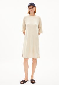 Beige knitted dress with short sleeves, side slits, and a loose fit. Paired with black slide sandals and a navy baseball cap.