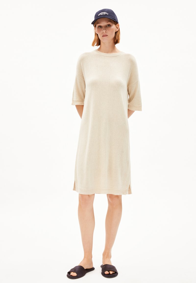 Beige knitted dress with short sleeves, side slits, and a loose fit. Paired with black slide sandals and a navy baseball cap.