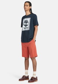 Camiseta de manga corta de Timberland en color navy con logo de árbol blanco, combinada con pantalones cortos color coral, zapatos de barco en crema y marrón, y calcetines rayados en crema.