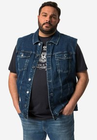 Dunkelblaues Denim-Westen, mit Knopfverschluss, zwei Brusttaschen und lockerer Passform, getragen über einem schwarzen T-Shirt und blauen Jeans.