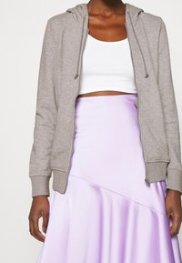 Sweat à capuche gris zippé avec cordons, présentant une texture lisse. Associé à un top court blanc et une jupe en satin violet clair.