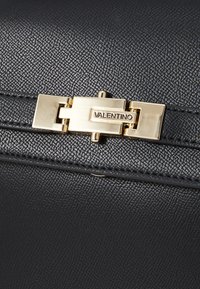 Borsa a tracolla in pelle nera con superficie testurizzata, chiusura in metallo dorato e logo "Valentino" impresso sulla chiusura.