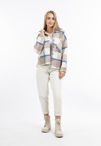 Geknoopte wollen blend jas met een beige en blauwe ruitpatroon, voorzien van een ronde kraag en twee voorzakken. Gecombineerd met lichte broeken en beige laarzen.