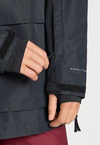 Črna vodoodporna jakna s kufrom na zadrgo in nastavljivim Velcro trakom, teksturirana tkanina in zategovalna vrvica na robu.