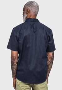 Kurzarmhemd in Marineblau, mit glatter Textur, klassischem Kragen und dezenten Nähten auf der Rückseite, kombiniert mit hellen Shorts.