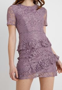 Krajková šaty v světle fialové barvě s kulatým výstřihem, krátkými rukávy, peplum pasem, vrstevnatými volány a květinovým vzorem po celém povrchu.