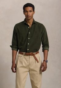 Tume roheline nööpidega särk, puuvillane kangas; beežid volangpüksid; kudumine pruun vöö; igapäevane käekell randmel; ühtlane taust.