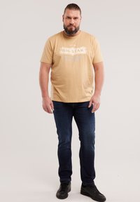 Mężczyzna z brodą i związanymi do tyłu włosami, ubrany w beżowy t-shirt z grafiką, ciemnoniebieskie dżinsy i czarne trampki, stoi na tle jednolitego tła.