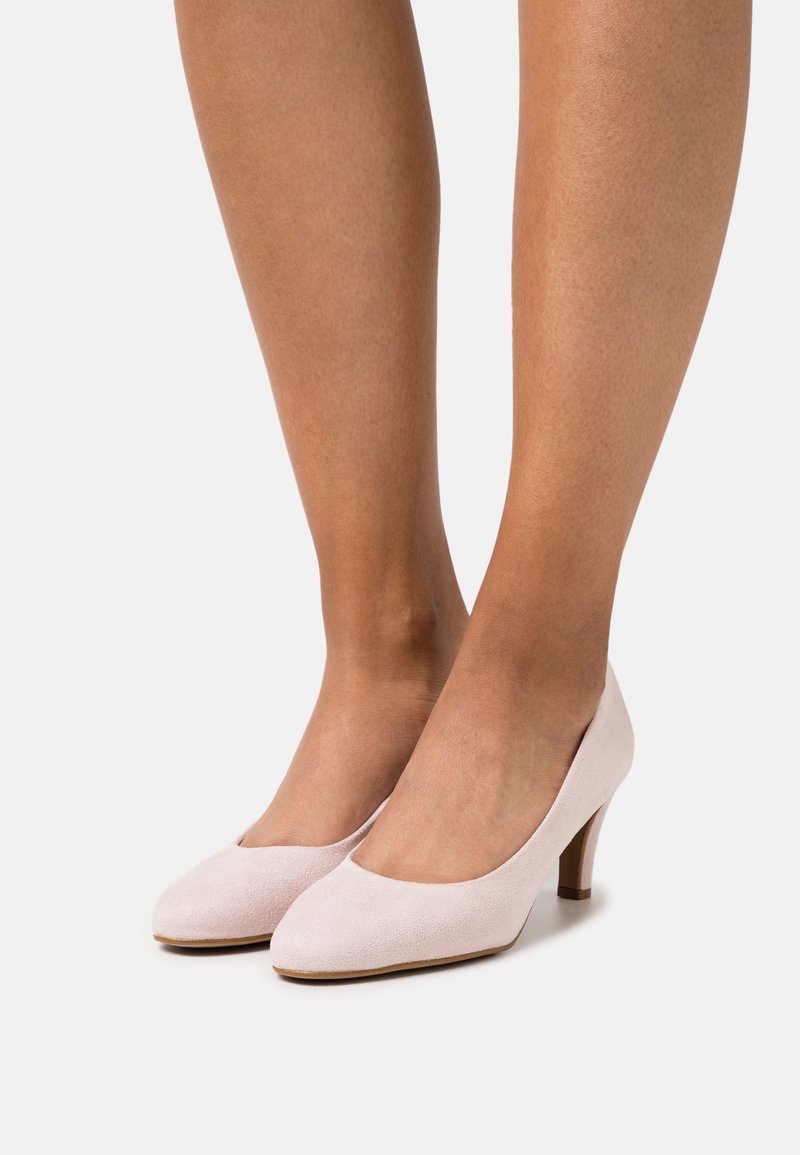 Paire d'escarpins en daim rose clair à talons moyens, portés sur des pieds à teint moyen, vus de face et de côté sur un fond uni.