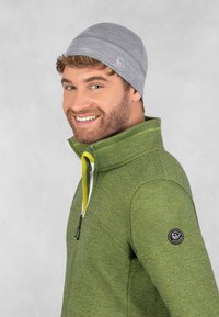 Bonnet gris avec une finition texturée et logo, associé à une veste en polaire verte zippée avec un badge circulaire noir et des accents jaunes.