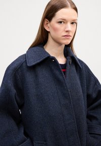 Cappotto in misto lana blu navy con grande colletto, bottoni frontali e tasche laterali. Presenta una finitura testurizzata e una vestibilità leggermente oversize.