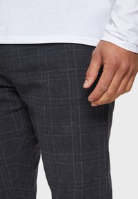 Plaid pants in dark gray fabric, featuring a textured weave and subtle checks. Hand resting near waist, with a white long-sleeve shirt visible.