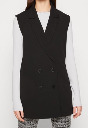 Femme portant un blazer noir sans manches à double boutonnage sur un haut blanc à manches longues et un pantalon à motifs noir et blanc, cadré au niveau du menton et des cuisses.