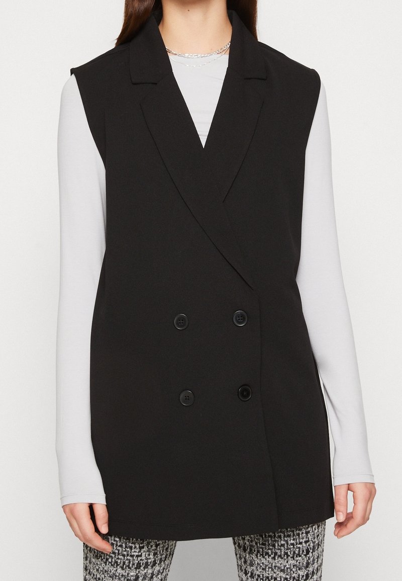 Femme portant un blazer noir sans manches à double boutonnage sur un haut blanc à manches longues et un pantalon à motifs noir et blanc, cadré au niveau du menton et des cuisses.