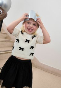 Lächelndes Kind in einem weißen Pullover mit schwarzen Schleifen und einem schwarzen Rock, das eine silberne Papierspur auf dem Kopf hält, in der Nähe einer Treppe.