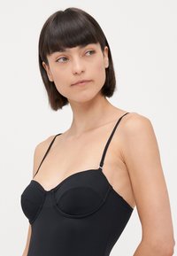 Juodas bodysuit'as su vieliniais puodeliais, reguliuojamomis plonomis petnešomis ir lygia matinė tekstūra. Yra formuoti puodeliai ir prigludusi siluetas.